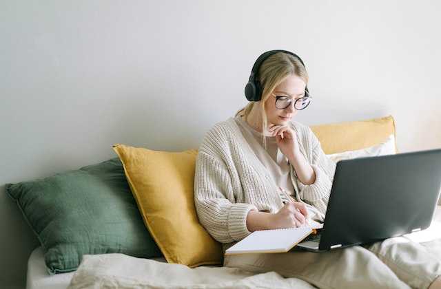Девушка сидит в наушниках, смотрит в ноутбук и записывает в тетрадь, как стать блогером и зарабатывать деньги