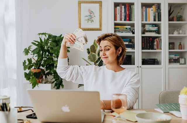 Женщина нашла способ, как заработать денег без вложений, поэтому держит в руке стопку купюр, сидя за компьютером