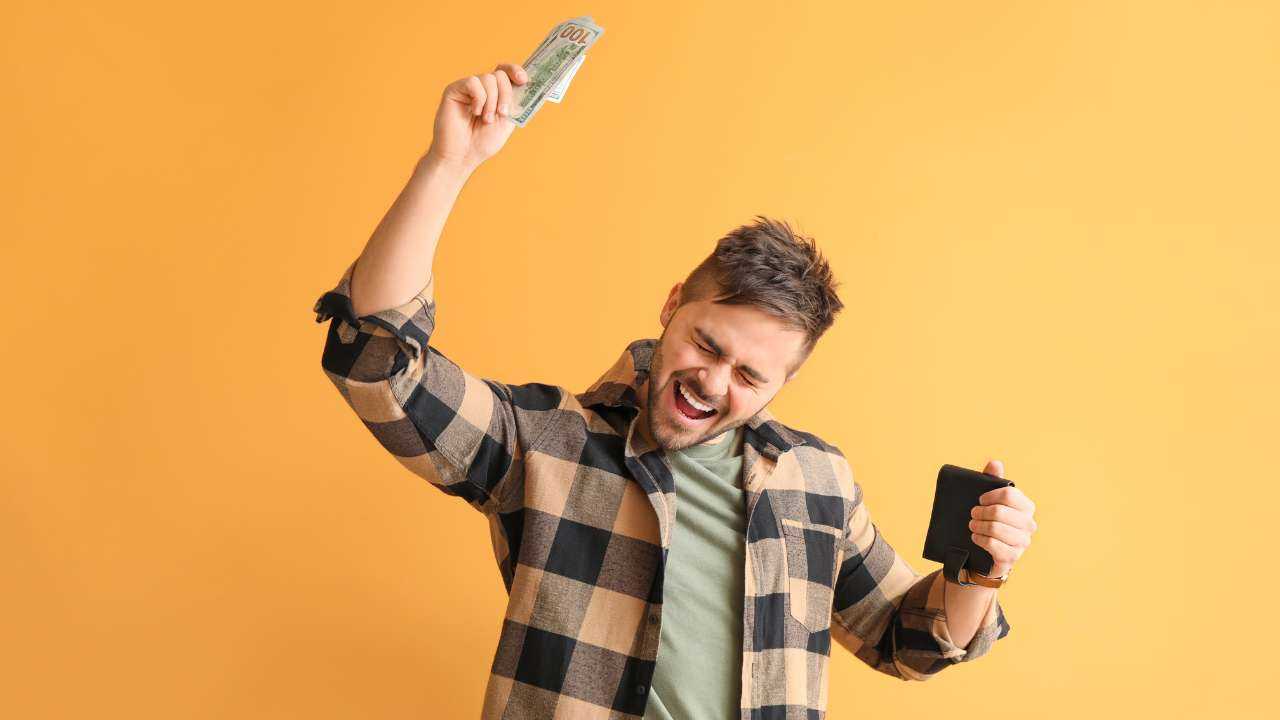 Молодой человек в клетчатой рубашке радостно машет купюрой на фоне желтой стены, иллюстрируя влияние денег на счастье