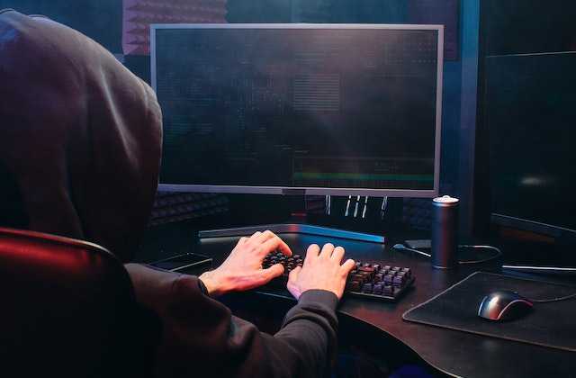 Мужчина в черном капюшоне сидит за компьютерами и программирует код, который помогает переводить деньги мошенникам