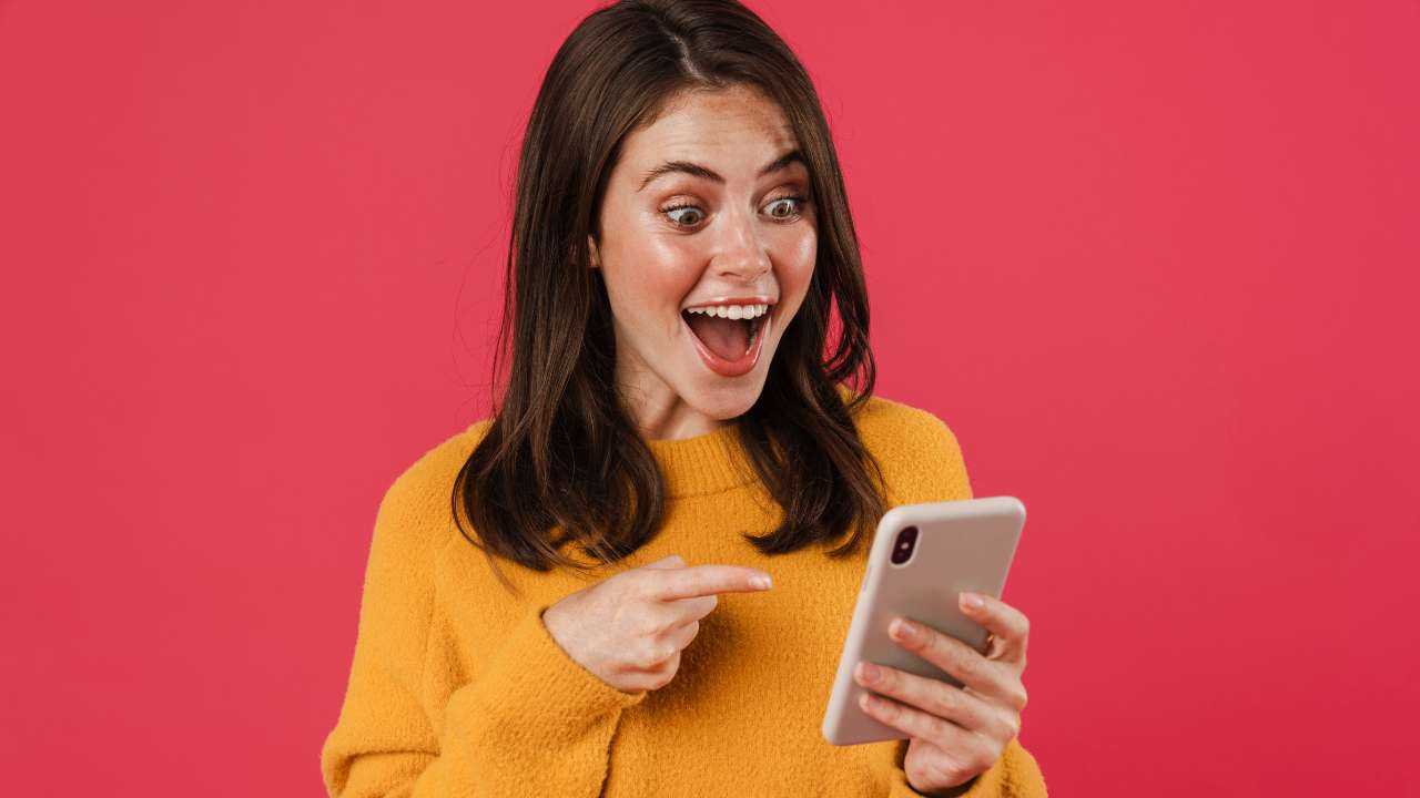 Tumšmate dzeltenajā džemperī priecājas un ar pirkstu rāda uz telefona ekrānu, uz kura ir atvērta mājaslapa MyCredy