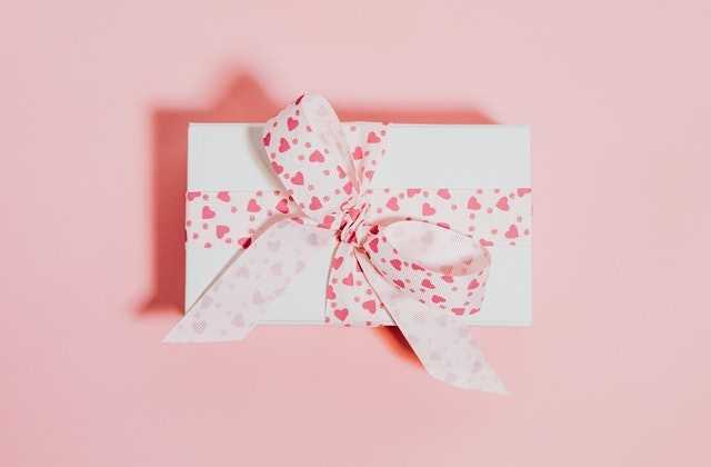 На розовом фоне лежит белый конверт, перевязанный лентой с сердечками, в котором внутри лежат деньги, подаренные на свадьбу